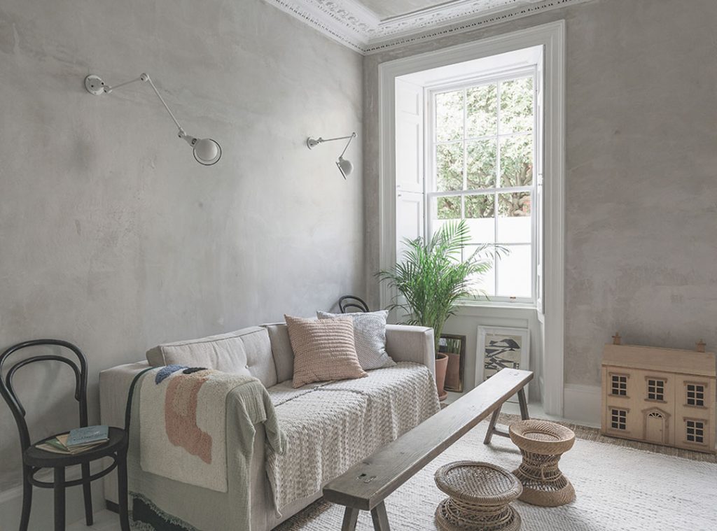 A corner of the neutral living room with a cream sofa, long bench coffee table, and angle poise wall lamps. A wooden dollhouse sits on the floor. 