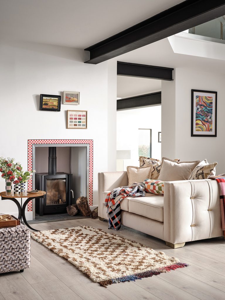 A neutral beige sofa sits beside a fireplace with a red and white chequerboard border. The walls of this living room are white with black beams overhead.