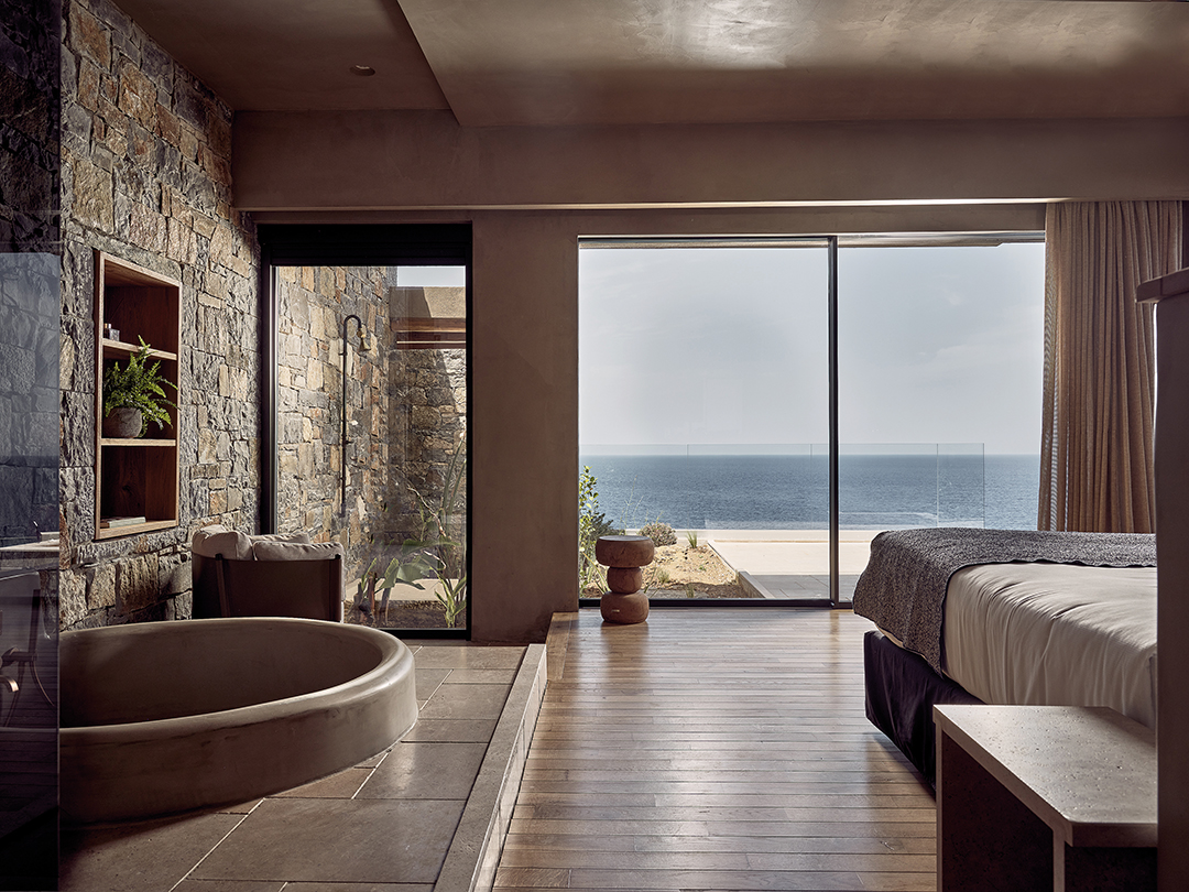 Definitely a bucket list item. This neutrally-decorated hotel room has sliding doors looking out onto sandy beaches and the ocean. To the left of the bed is a sunken, round bath and through another window you can see an outdoor shower.