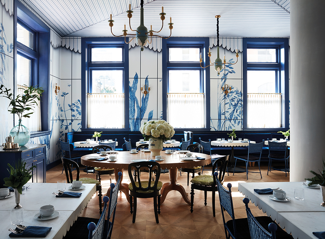 An incredible dining room finished in blue and white. Chandeliers overhead, white flowers in vases on the tables, scallop-edged tables in the foreground