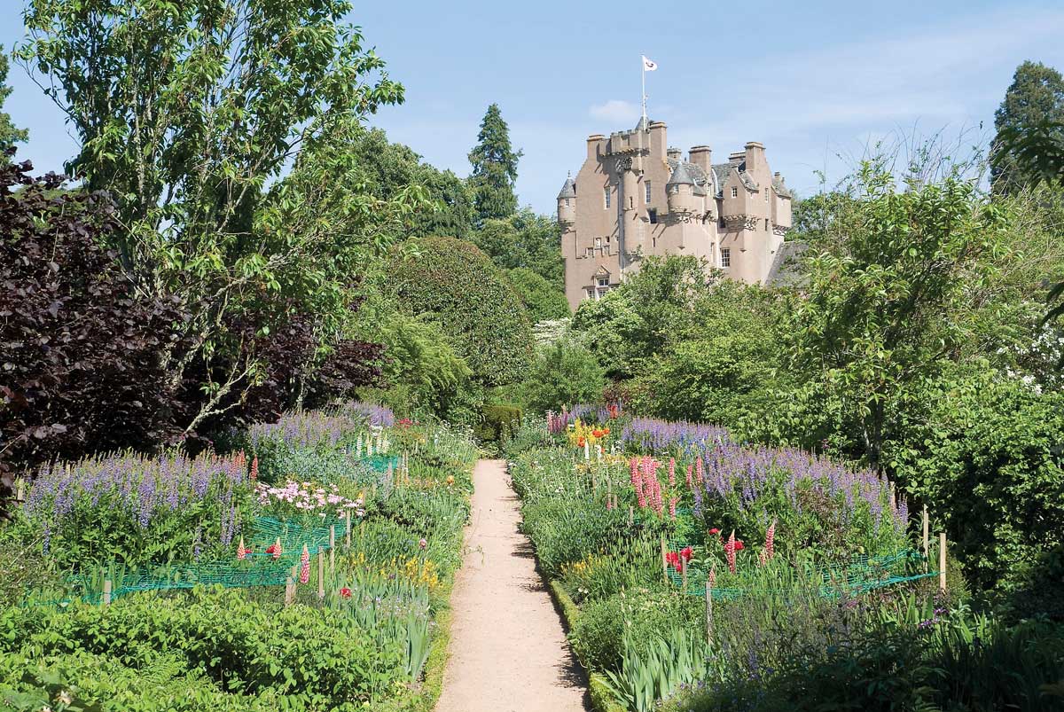 Crathes Castle, one of the National Trust for Scotland's impressive gardens. A light coloured castle with a flag sits in the background, colourful flowers take up the foreground.