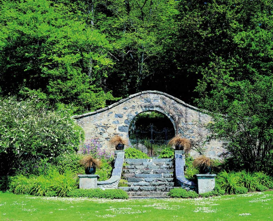 A round iron gate in a stone wall surrounded by greenery
