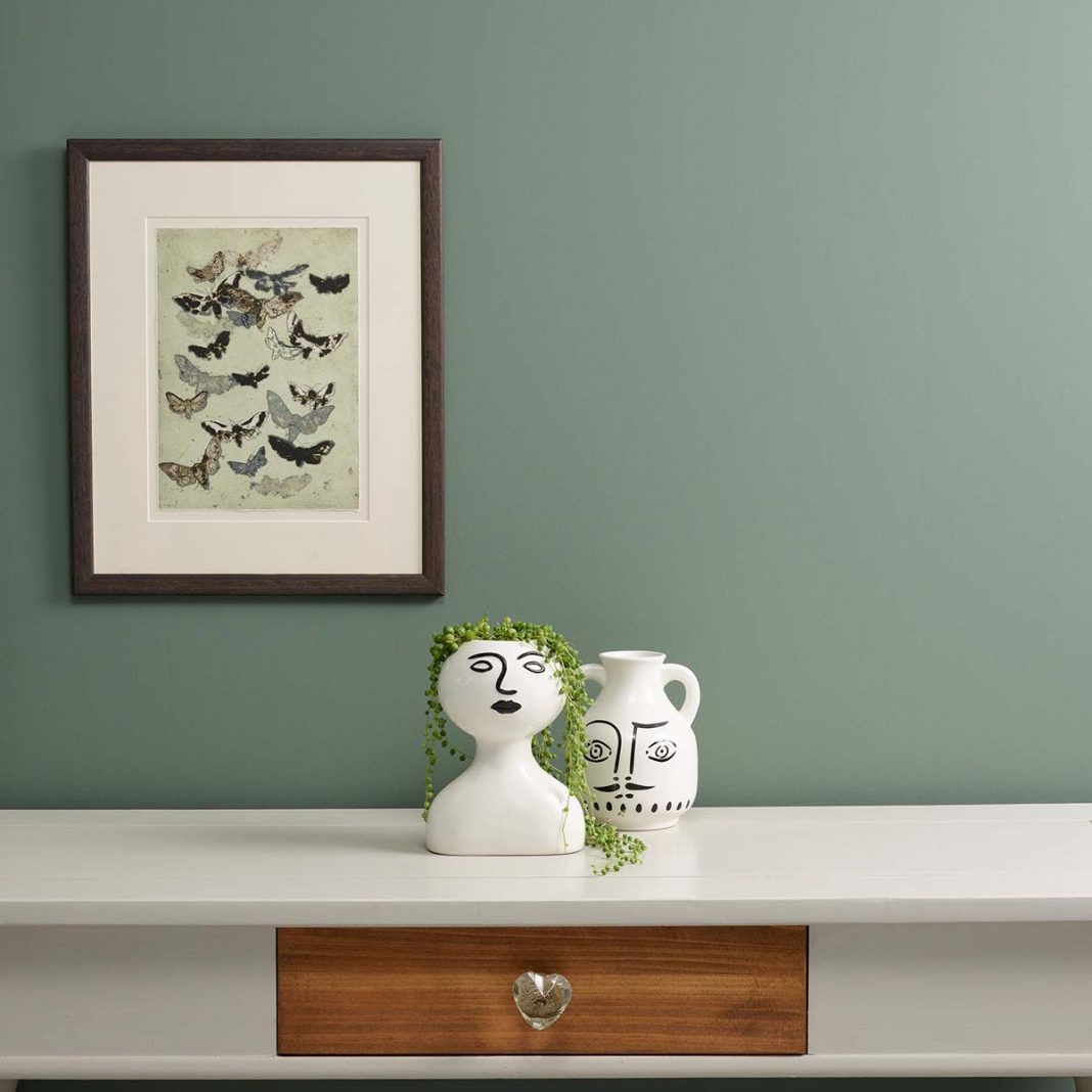 Wall painted in 'Steaming Green' by Frenchic Paint. There is a picture of butterflies on the wall and below that a table with two vases.