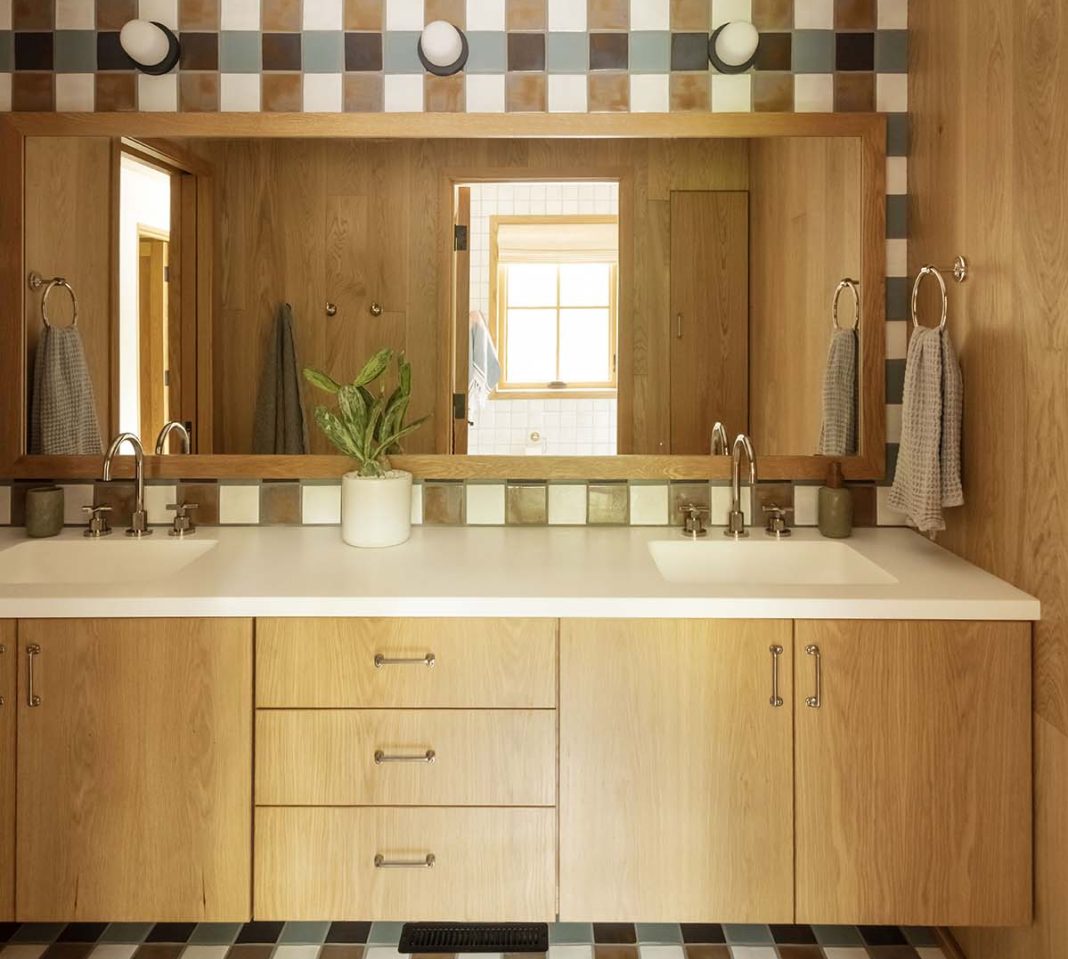 A bathroom with double sink and large mirror. The back wall and floor are tiled in a plaid pattern in shades of ivory, terracotta and blue. The cabinetry and other walls are covered in plain sawn white oak