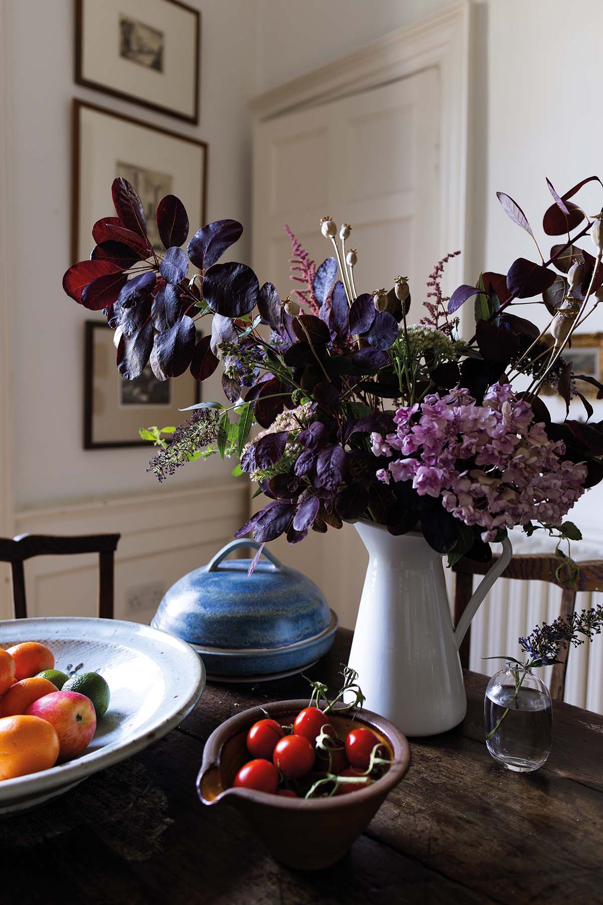 a closeup of flowers in a jug, a fruit bowl, tomatoes and a pot on a dining table