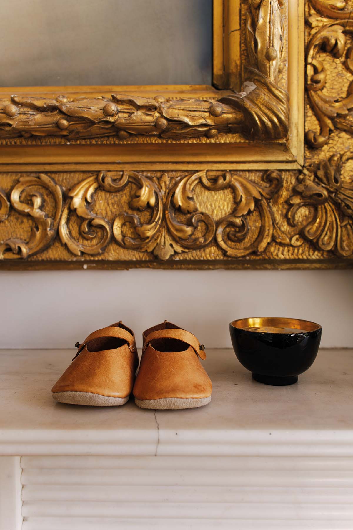 a pair of small brown shoes and a small dish sit on a mantlepiece under a gold framed mirror
