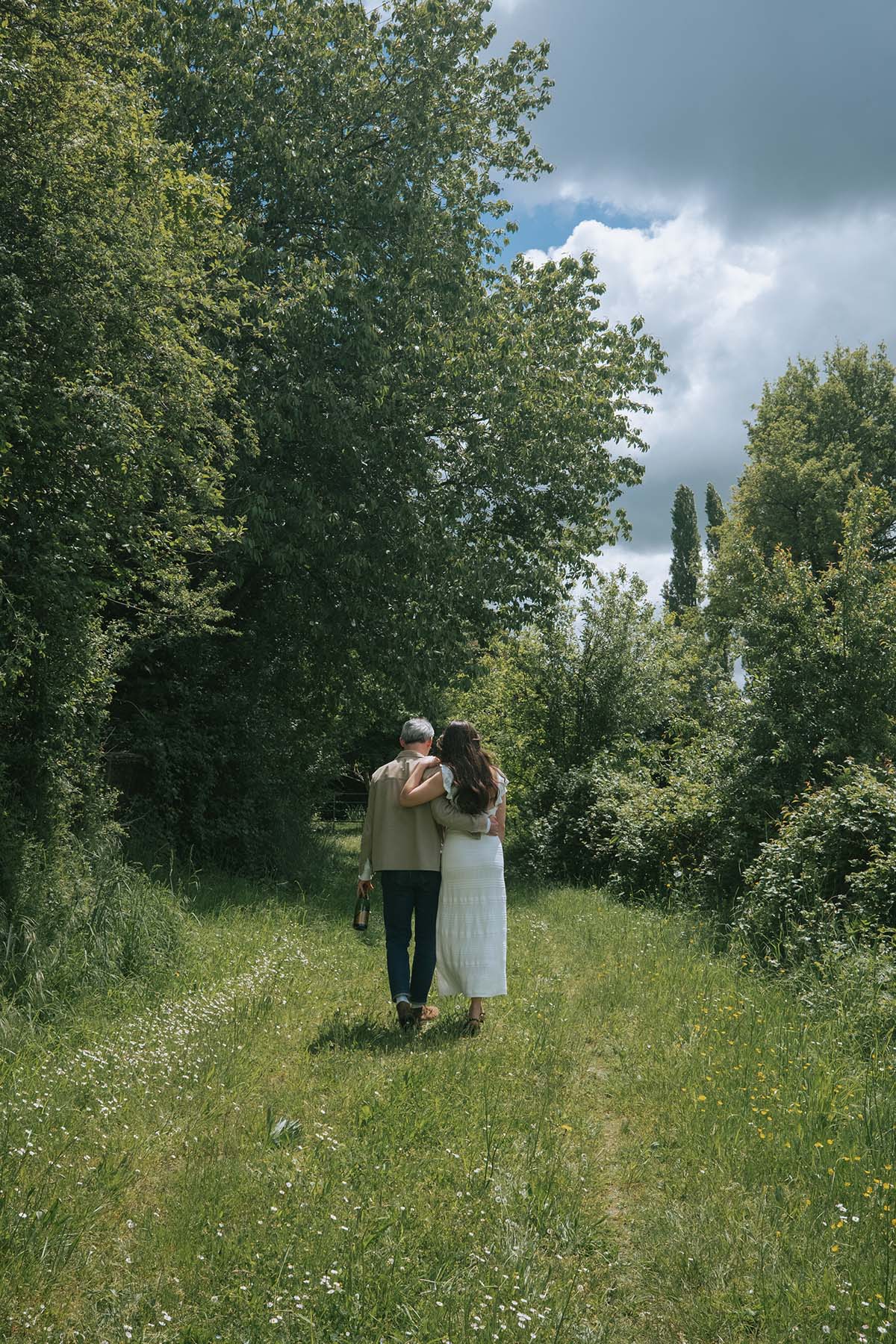 A couple embraces while walking away from the camera along an overgrown track