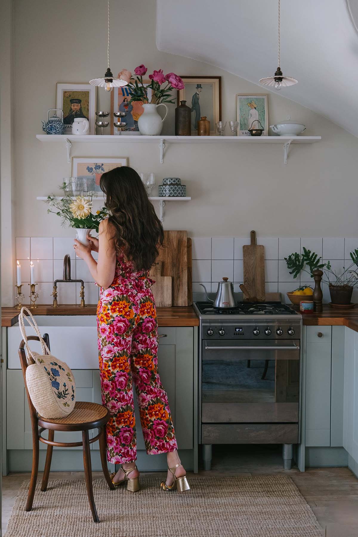 a woman in a floral jumpsuit stand ins her kitchen, holding a vase