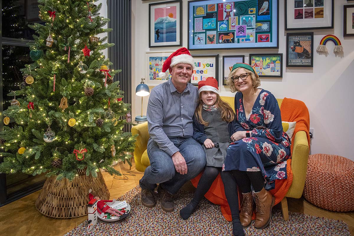 a dad, mum and daughter sit on a sofa next to a Christmas tree