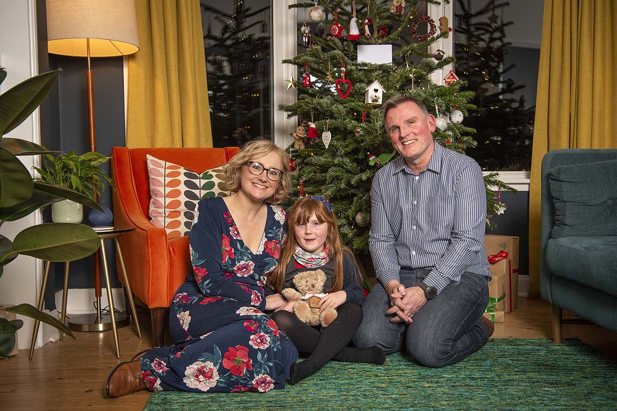 A mum, dad and young daughter sit on the floor in front of a Christmas tree in a living room