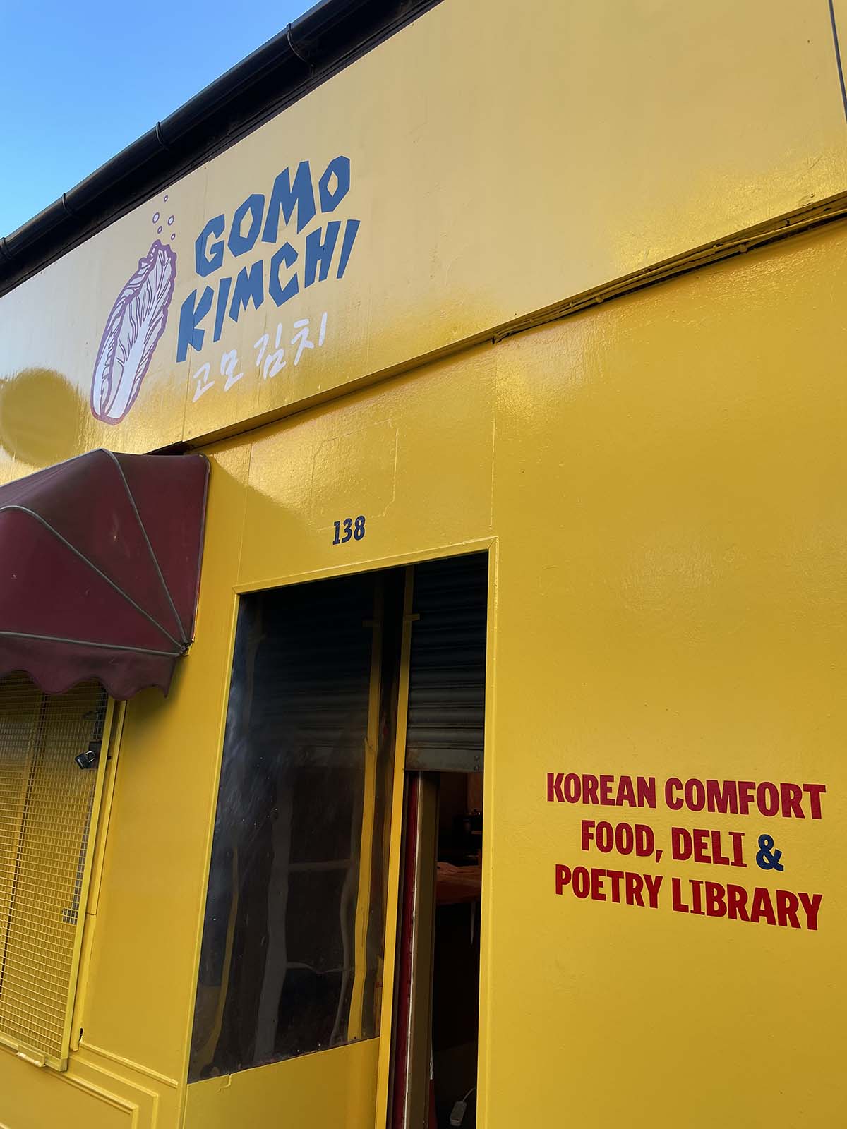 a bright yellow store front with 'Gomo Kimchi' written at the top and 'Korean Comfort Food, Deli & Poetry Library" written by the door