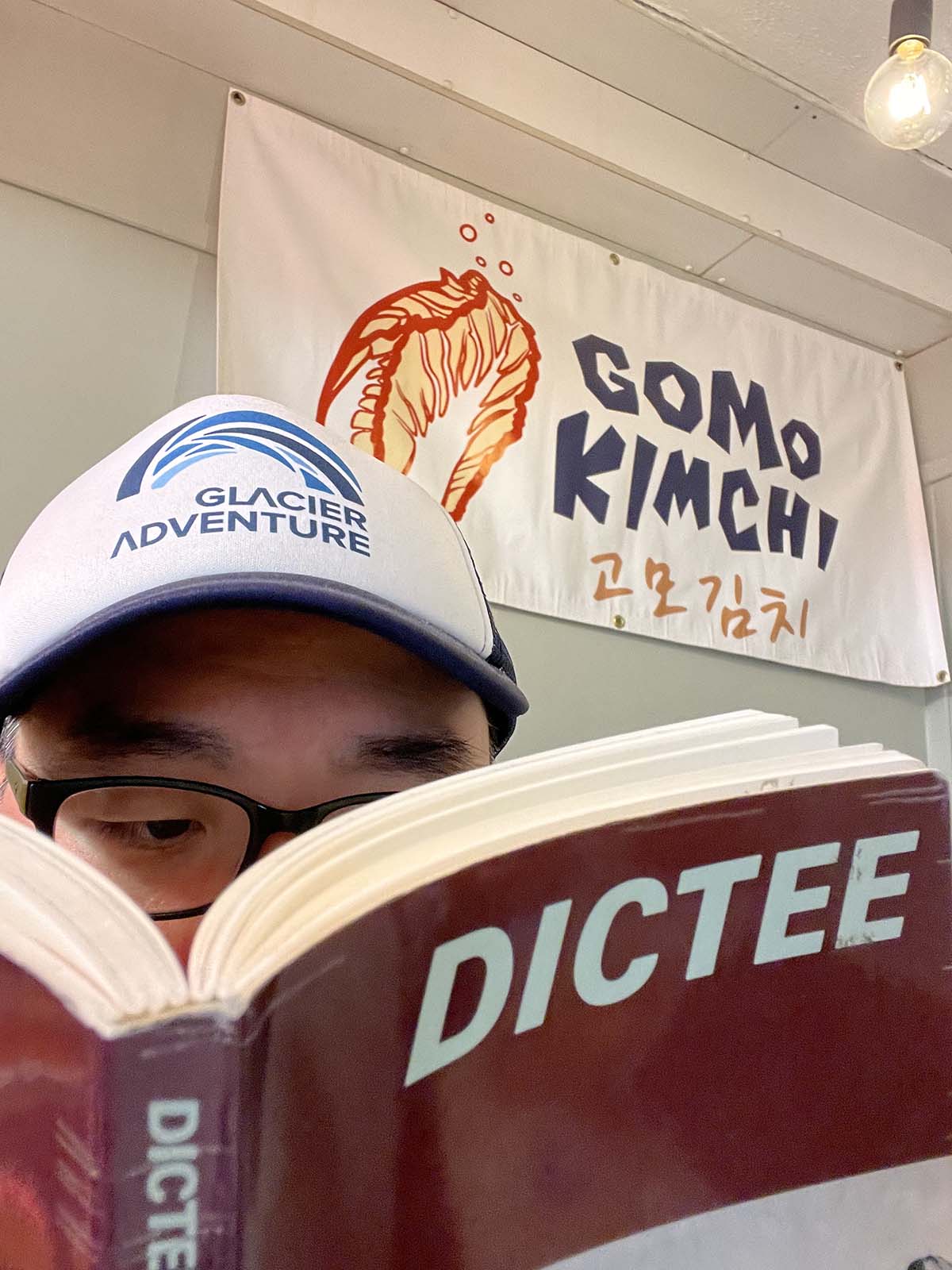 a man wearing a baseball cap and glasses reads a book, underneath a banner that says 'Gomo Kimchi'