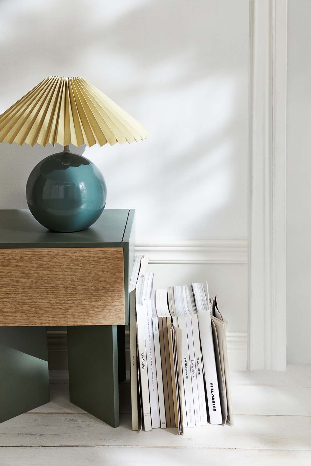 a lamp with a neutral pleated lampshade and a green base sits on a low table with book propped against it