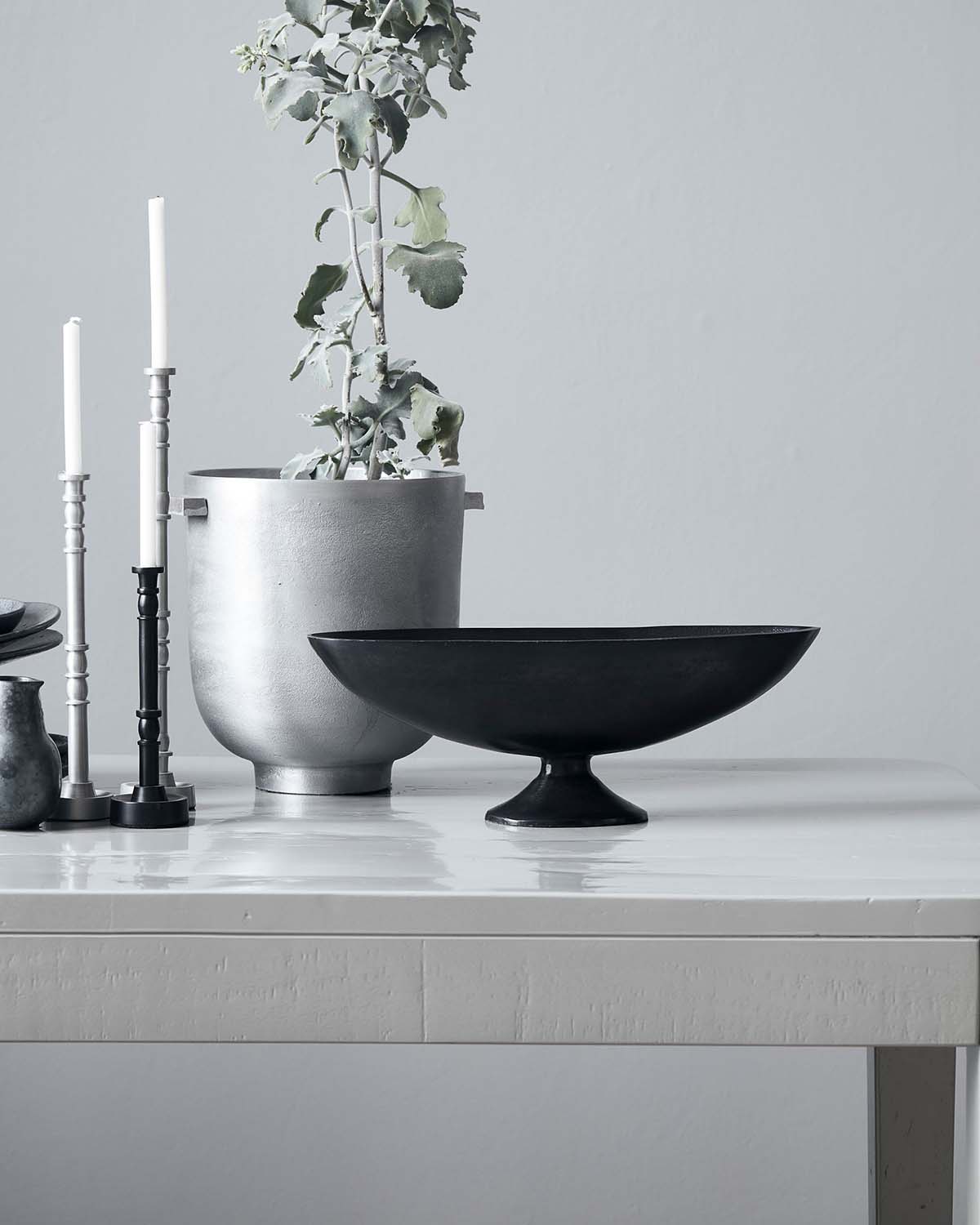 A black bowl sits on a sideboard with a silver plant pot and candlestick holders next to it