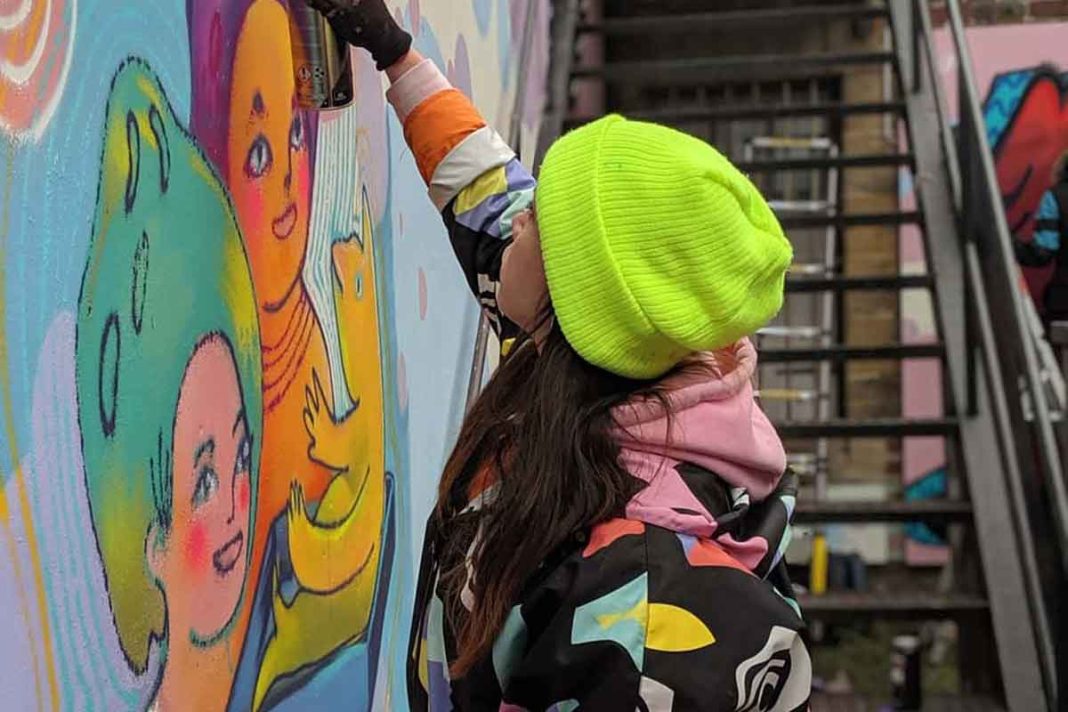 A woman wearing a bright green beanie and a bright, patterned coat spray paints a wall