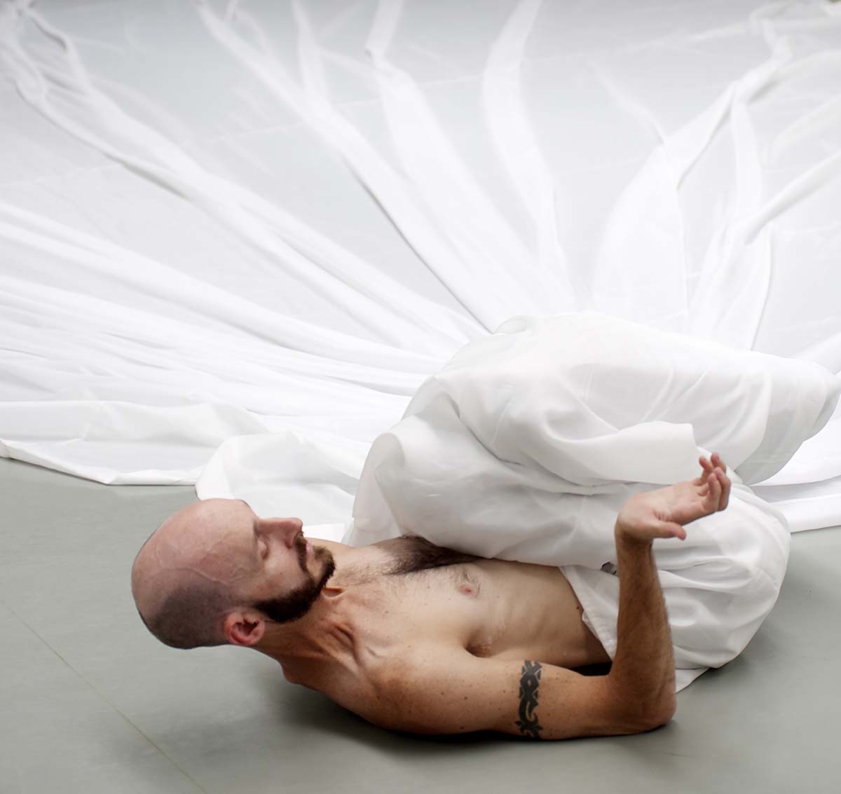 A man lies with his legs curled into his chest, a white sheet covering them and splayed out behind him. His chest is bare and he has a tattoo on his right upper arm.