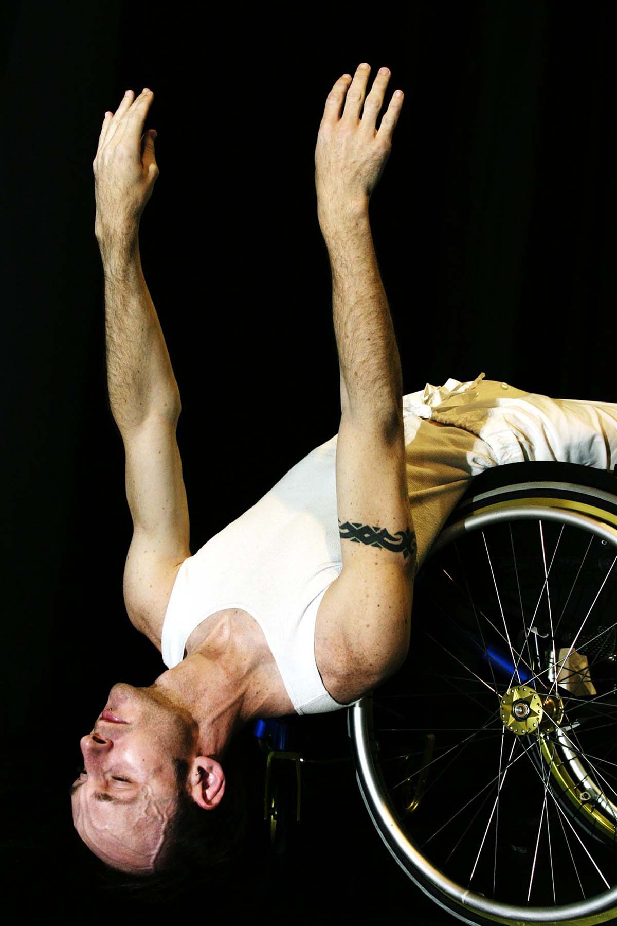 A man in a white vest and white trousers leans back over the wheels of a wheelchair, his arms stretched upwards