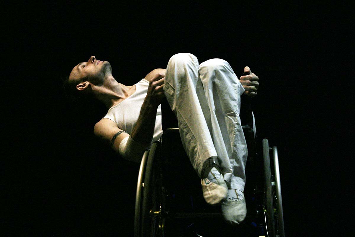 A man in a white vest and white trousers leans back in a wheelchair. He is shot from below