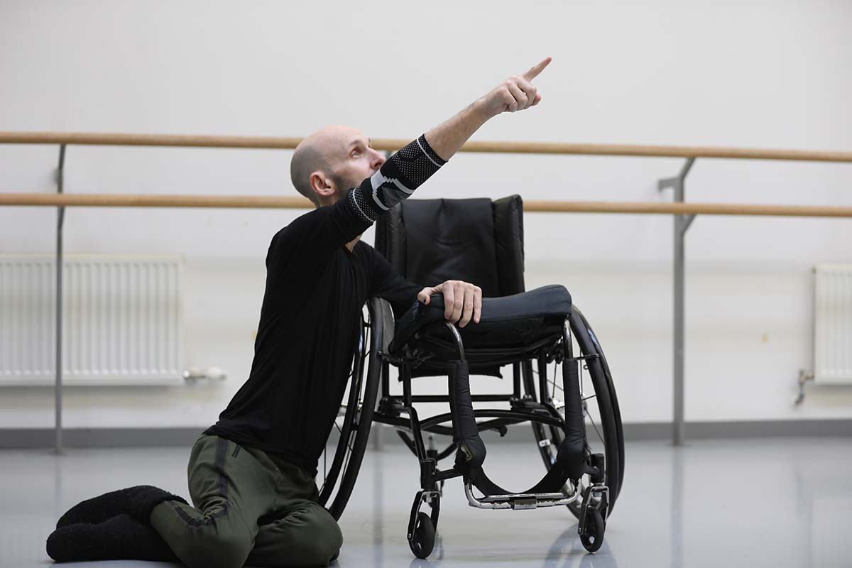 a man sits on the floor, leaning against a wheelchair and pointing up at something out of the frame