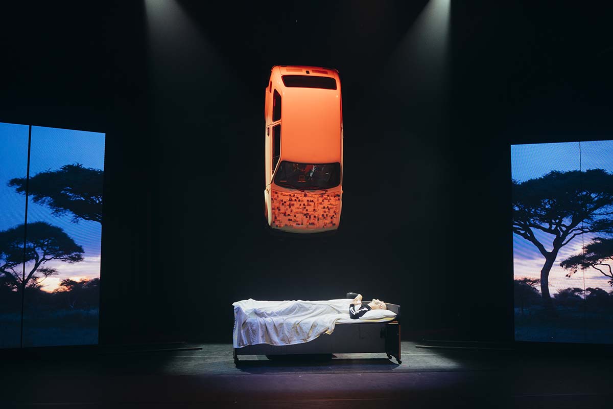 A stage with two LED screens showing a bush scene. Between the screens, a car hangs upside down and below the car is a man lying in a bed, covered by a sheet
