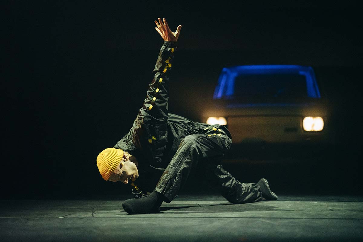 A man in a dark green tracksuit and yellow beanie crouches on the floor in a low lunge, one arm stretched upwards and behind him. A car with its headlights on is behind him