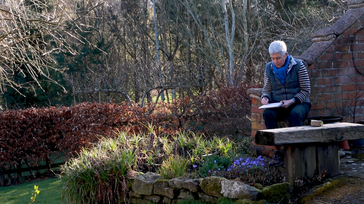 pauline burbidge in her garden with flowers around her sitting on a stone wall