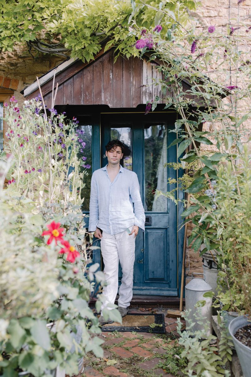 man stands in front of a blue door of a cabin surrounded by gardens